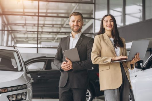Super sales team in dealership, two consultants or managers in elegant suits with laptop and tablet in arms in car dealership Super sales team in dealership, two consultants or managers in elegant suits with laptop and tablet in arms in car dealership